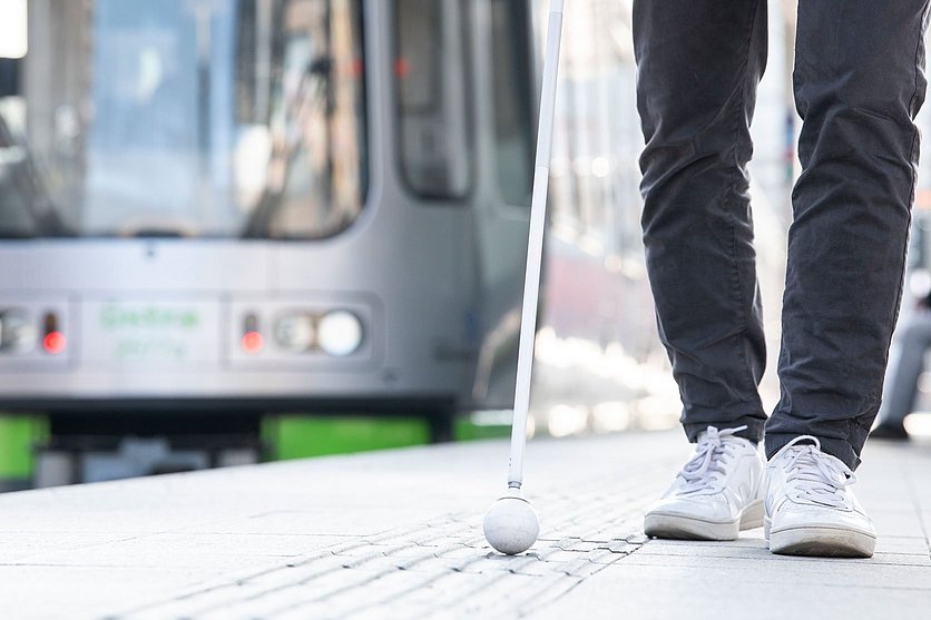 Bild von einer Person mit Blindenstock auf dem Bahnsteig mit einer Stadtbahn im Hintergrund 