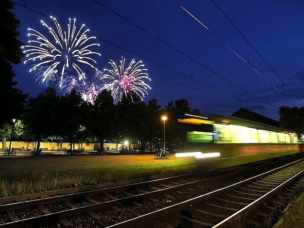 Eine ÜSTRA Stadtbahn fährt vorbei und im Hintergrund ist das Feuerwerk am Abendhimmel zu sehen.