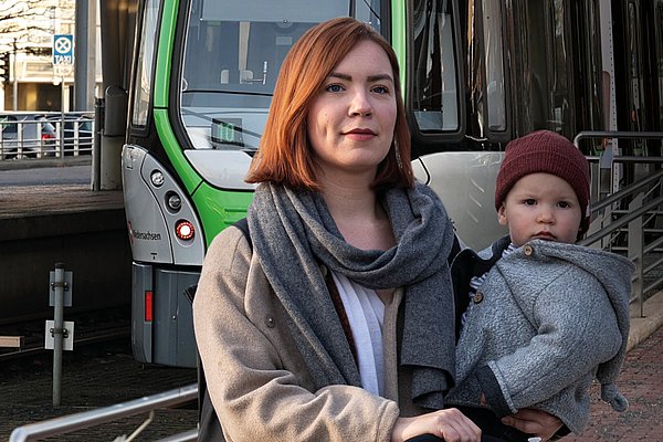 Titel des GVH journal 01.2022: Eine Frau hält ihr Kind auf dem Arm und schaut in die Kamera. Im Hintergrund fährt eine Stadtbahn ein.