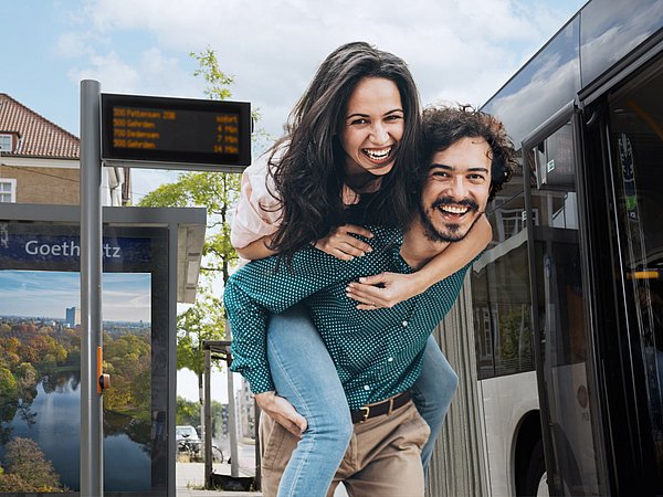 Bild von einem Pärchen an einer Bushaltestelle. Die Frau wird dabei vom Mann Huckepack genommen.