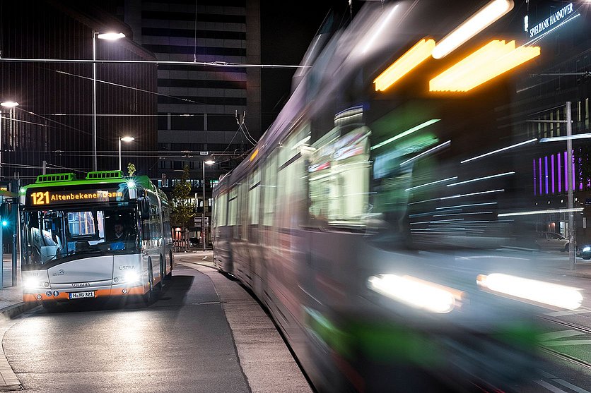 Bild von einem Bus, der nachts an einer Haltestelle hält, eine Stadtbahn fährt vorbei.