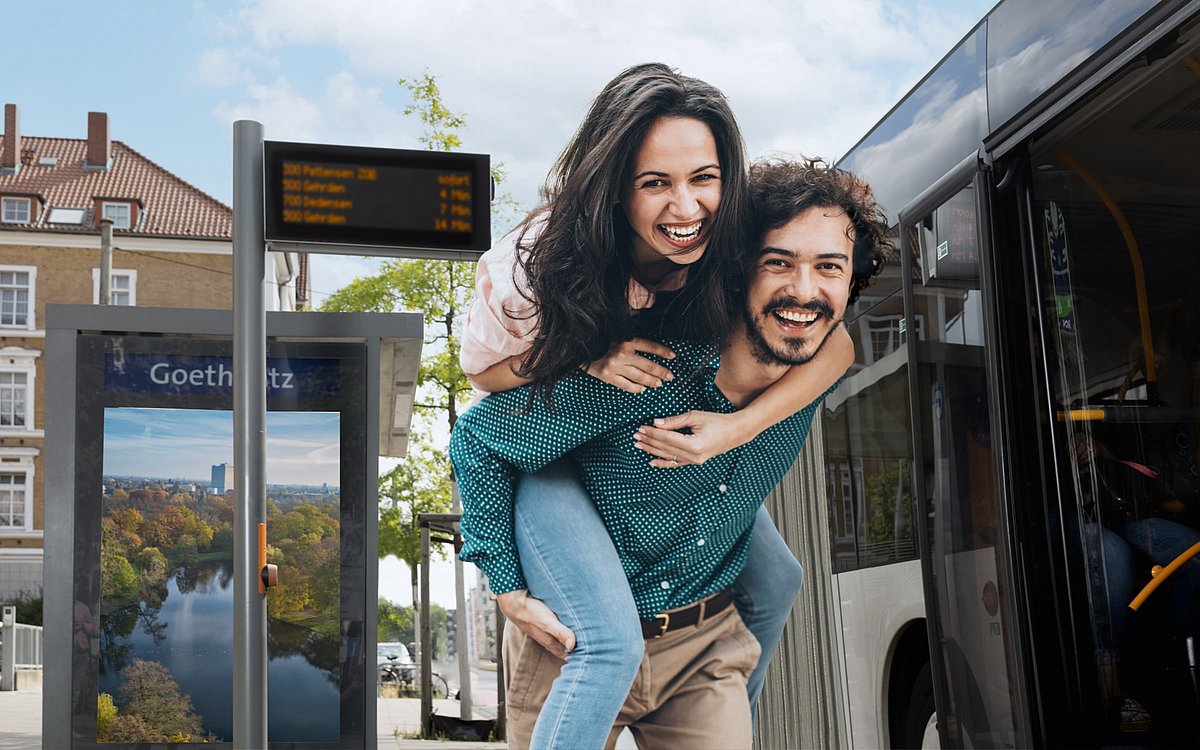 Bild von einem Pärchen an einer Bushaltestelle. Die Frau wird dabei vom Mann Huckepack genommen.